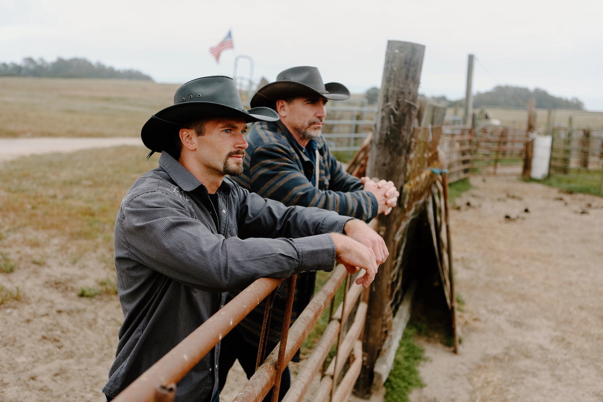 Two men wearing leather black cowboy hats leaning on the ranch's metal fence