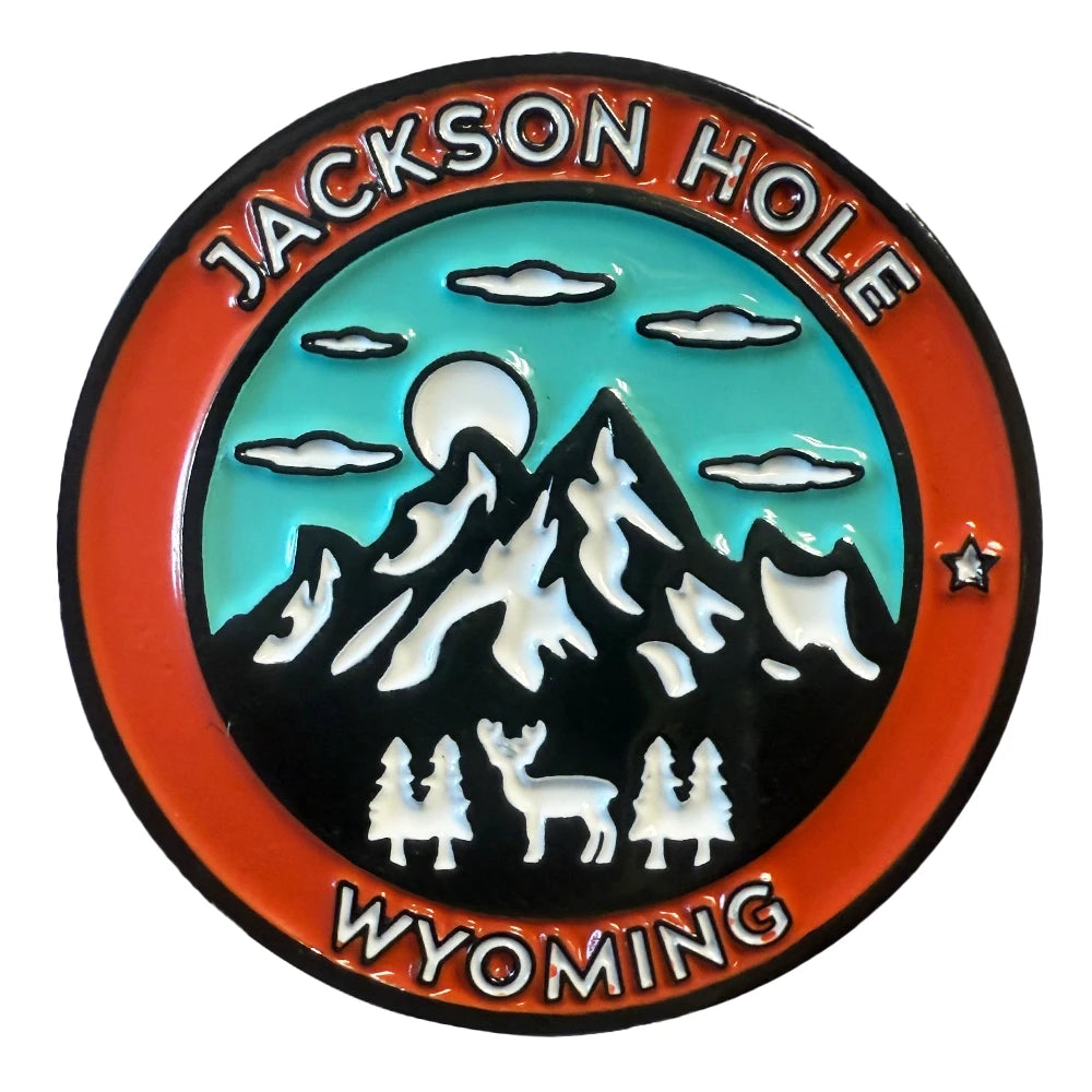 Round enamel pin featuring a Jackson Hole Wyoming mountain landscape with a deer, pine trees, and teal sky inside an orange border.