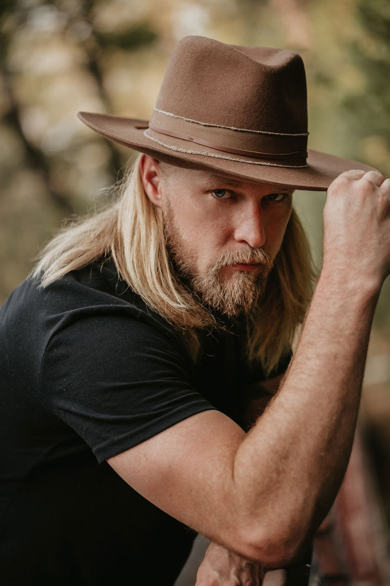 A man standing outdoors wearing a brown felt hat while holding it with right hand