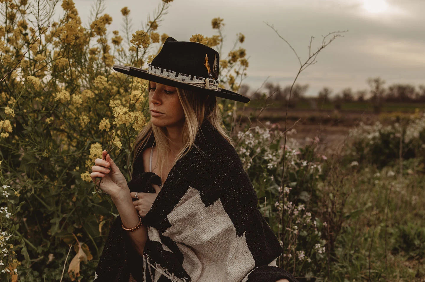 woman with a shawl looking at the flowers in the field wearing a black hat by american hat makers