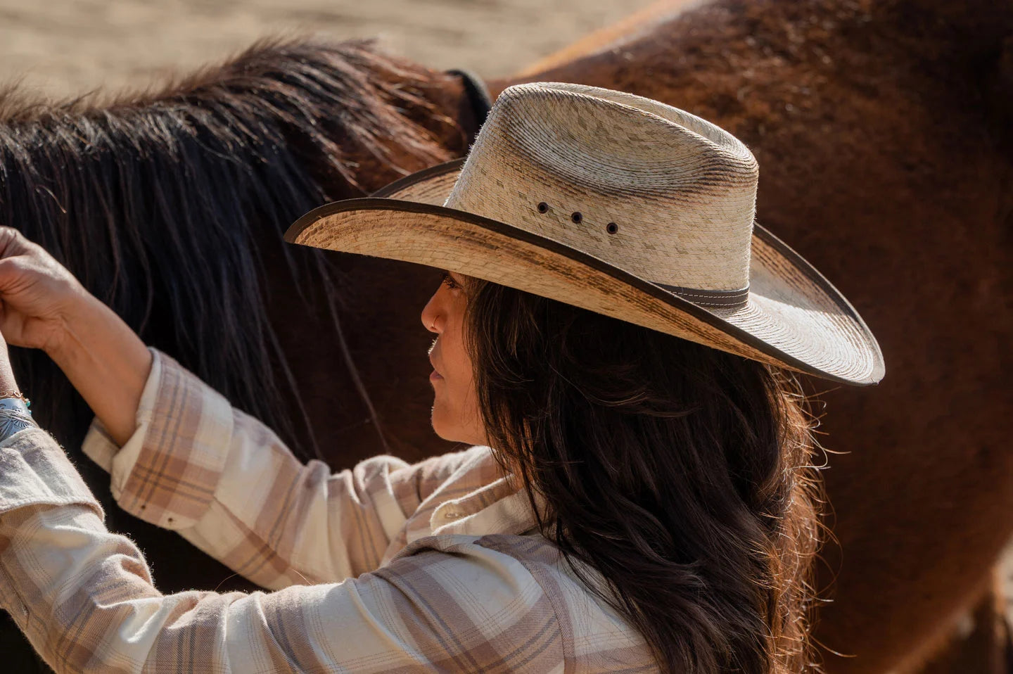 A woman tending to a horse wearing a brown checkered polo and a straw cowboy hat by American Hat Makers