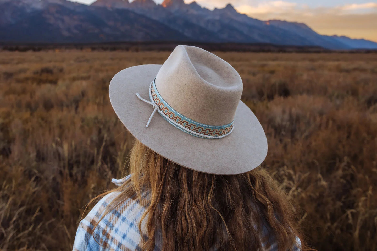 A woman in a field looking towards the mountains wearing a plaid shirt and a felt hat