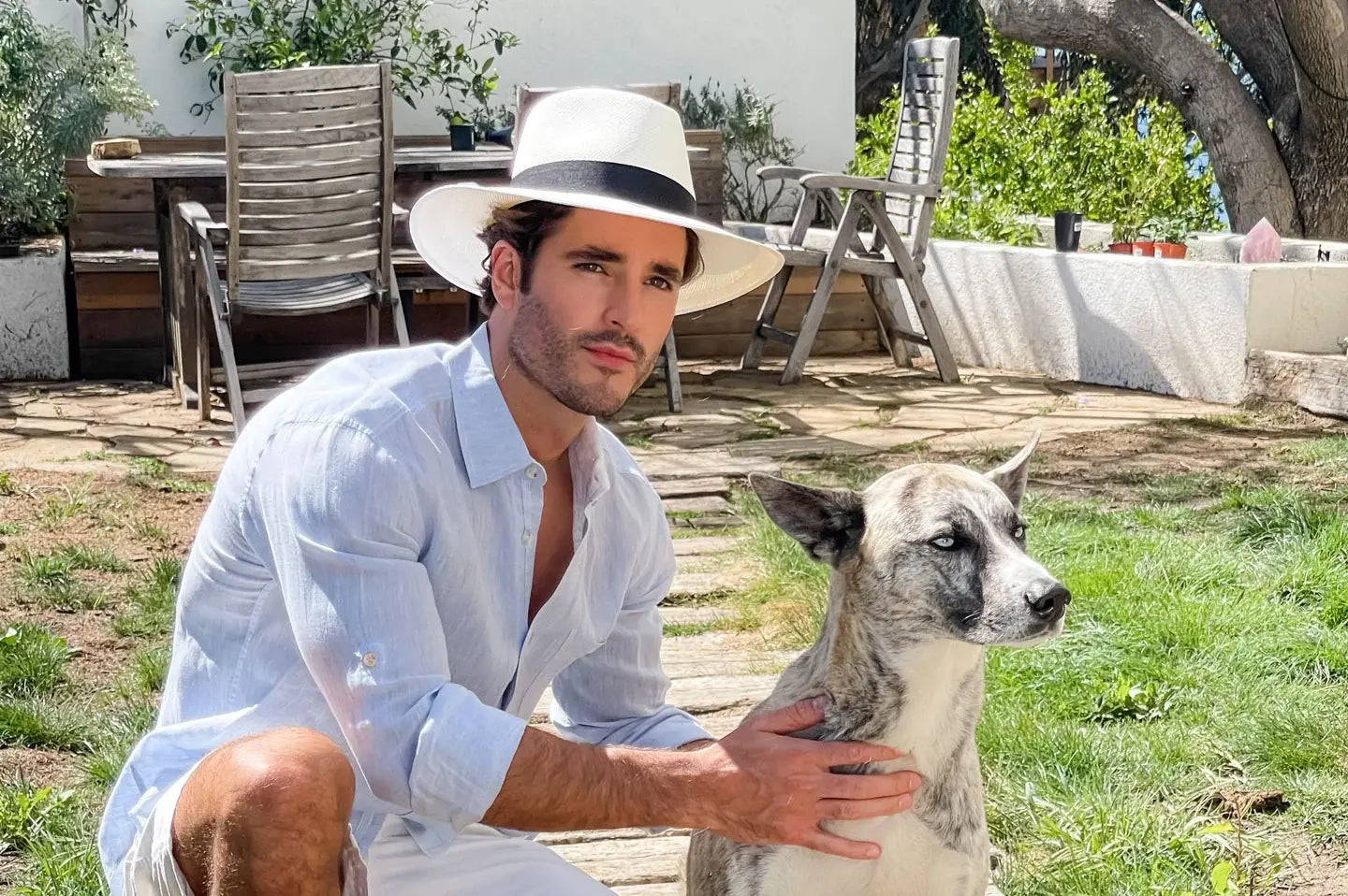 A man and his pet dog sitting on a garden wearing a white polo and sun hat 