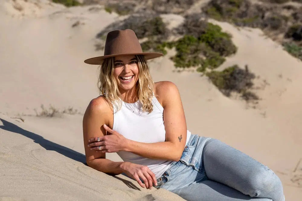 A woman lying on the sand wearing a white tank top, jeans and a brown felt hat
