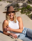 A woman lying on the sand wearing a white tank top, jeans and a brown felt hat