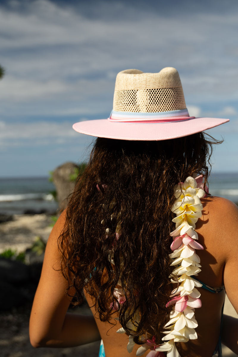 A woman on the beach with flowers around the neck wearing a pink straw sun hat