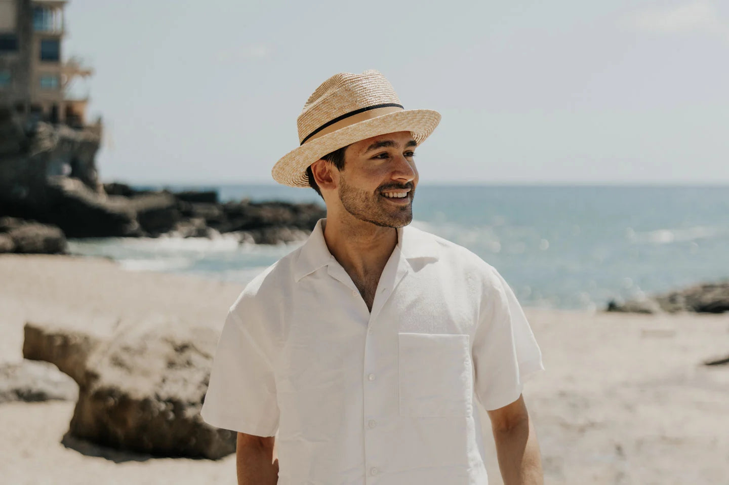 A man strolling on the beach wearing a white polo and a straw sun hat