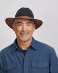 A man with a light stubble wearing a felt coal fedora hat and a gray button-up shirt.