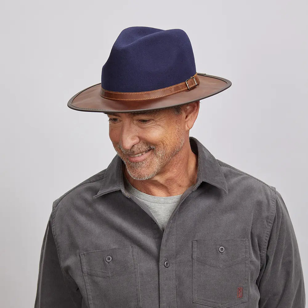 A man with a light stubble wearing a felt navy fedora hat and a gray button-up shirt.