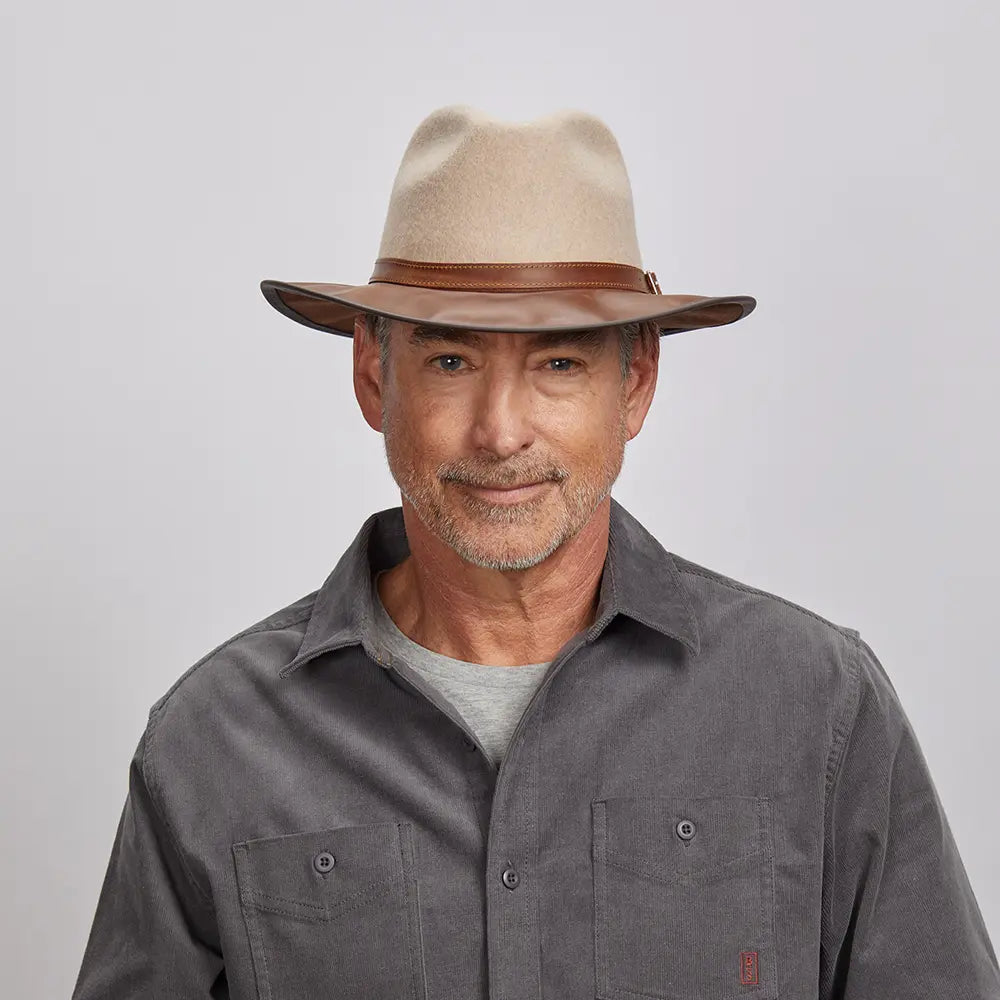 A man with a light stubble wearing an felt oatmeal fedora hat and a gray button-up shirt.