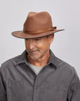 A man with a light stubble wearing a felt saddle fedora hat and a gray button-up shirt.