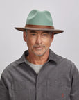 A man with a light stubble wearing a felt sage fedora hat and a gray button-up shirt.