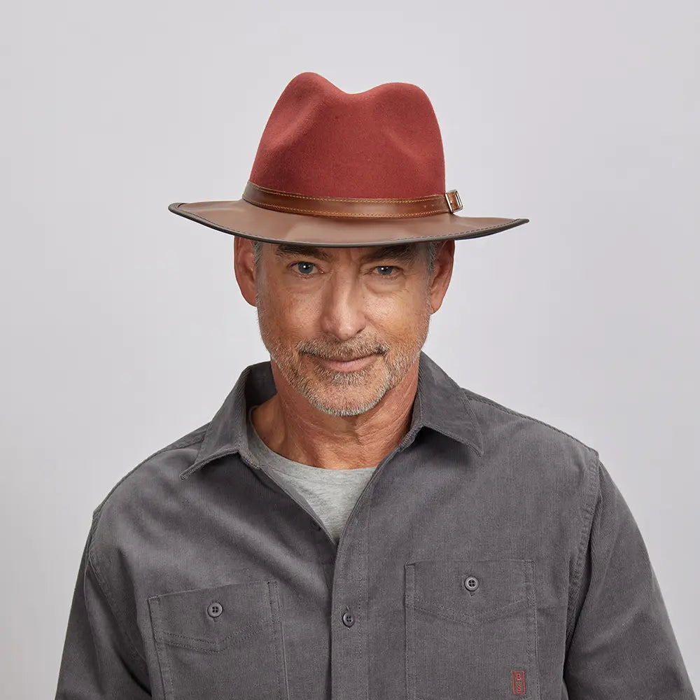 A man with a light stubble wearing a felt sangria fedora hat and a gray button-up shirt.