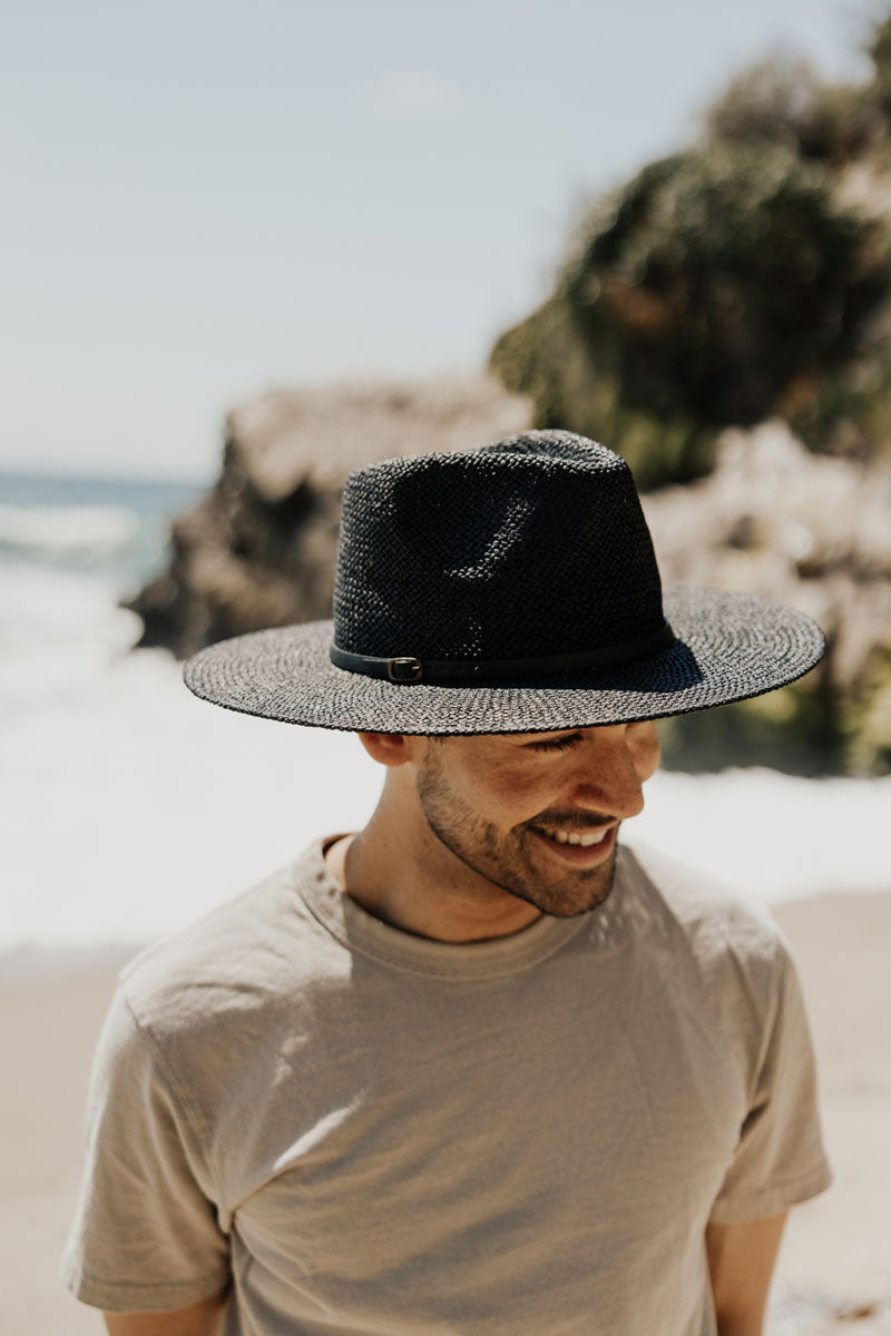 A man on the beach wearing a shirt and a black straw sun hat