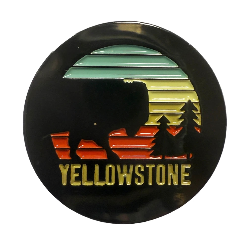 A circular enamel pin featuring a Yellowstone National Park design with a black silhouette of a bison, trees, and a sunset gradient in green, yellow, and orange, with 'YELLOWSTONE' written in gold at the bottom.