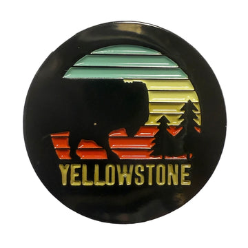 A circular enamel pin featuring a Yellowstone National Park design with a black silhouette of a bison, trees, and a sunset gradient in green, yellow, and orange, with 'YELLOWSTONE' written in gold at the bottom.