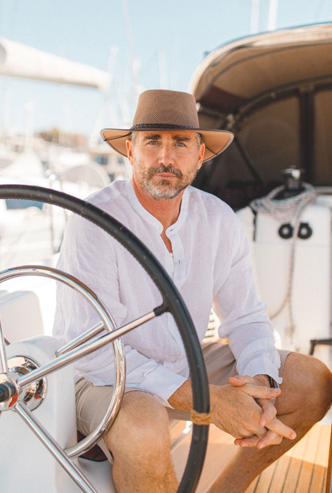 A man wearing a white top sitting behind a boat's wheel with this brown hat