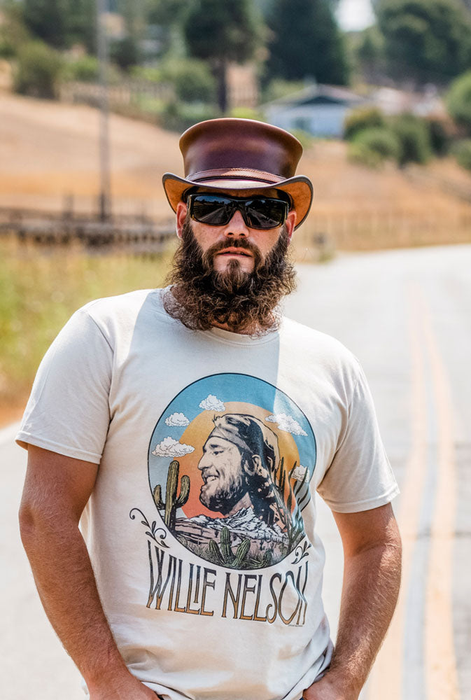 A bearded man in his Willie Nelson shirt and a brown top hat standing in the middle of the road