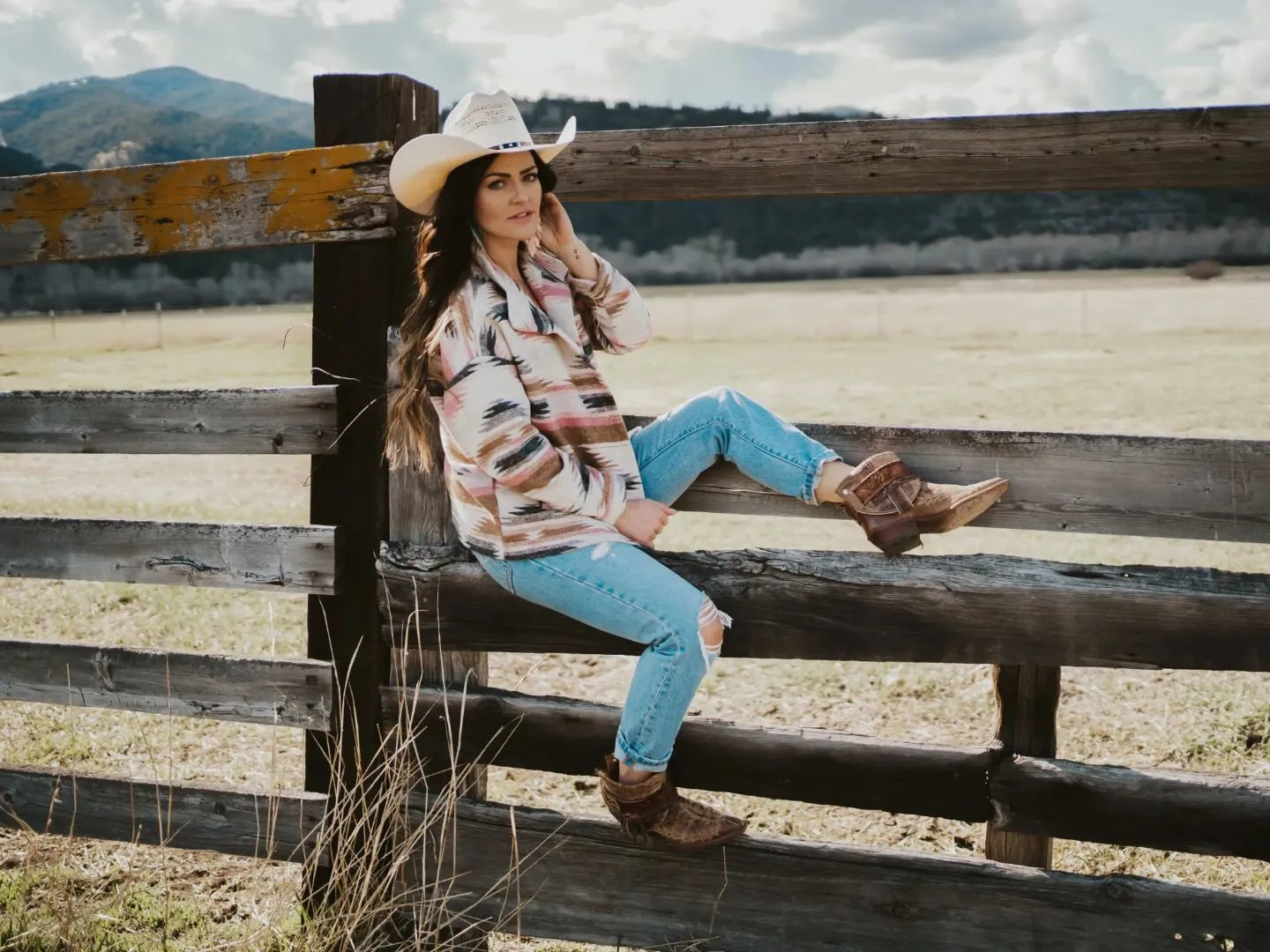 A woman sitting on a wooden fence wearing a boho jacket and cream cowboy hat