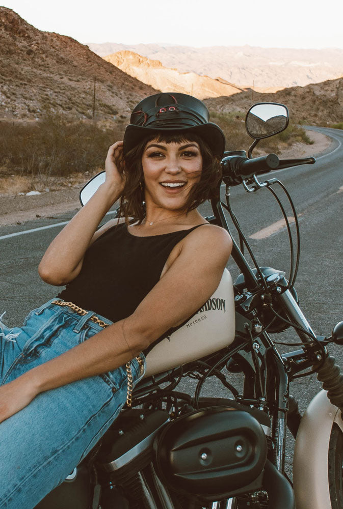A female biker leaning on his motorcycle wearing a black top, jeans and a black top hat