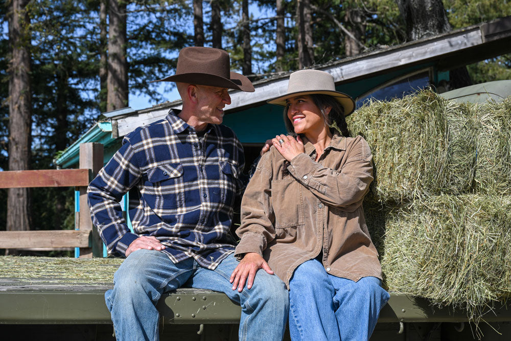 A couple sitting on a ledge beside a hay stack wearing yellowstone hats