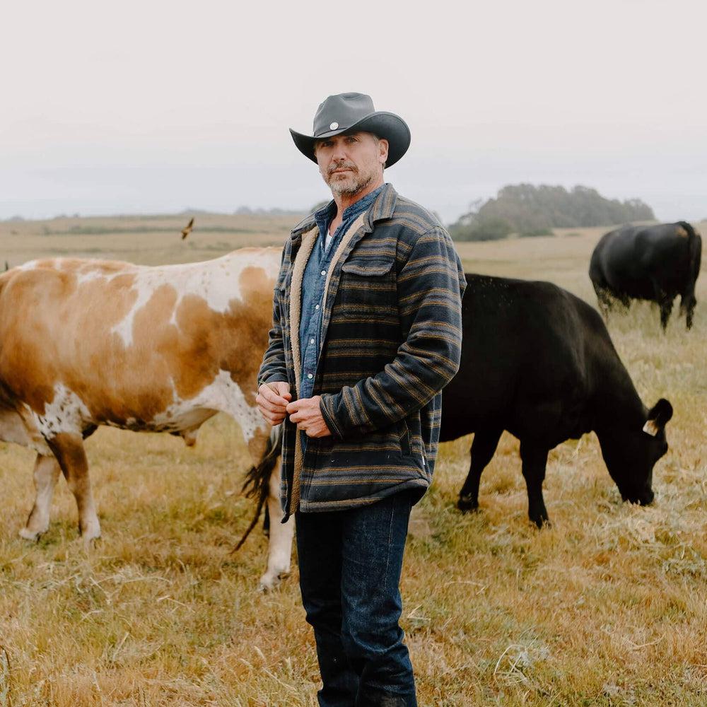 A man with cows behind him wearing Cyclone Black Leather Cowboy Hat with 3&quot; Brim and 4&quot; Crown