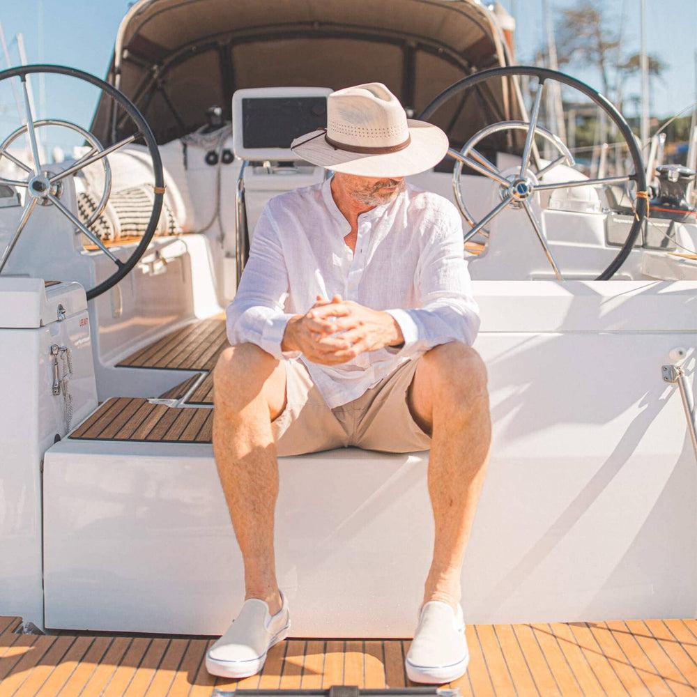 A man sitting on a boat wearing white shoes and a Barcelona Wide Brim Natural Straw Sun Hat 