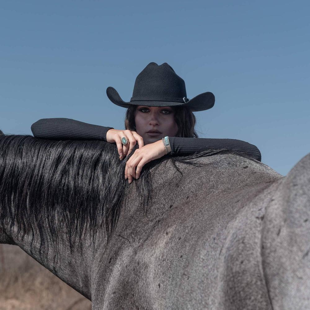 A woman standing behind a black horse and wearing a Felt Cowboy Hat 