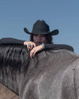 A woman standing behind a black horse and wearing a Felt Cowboy Hat 