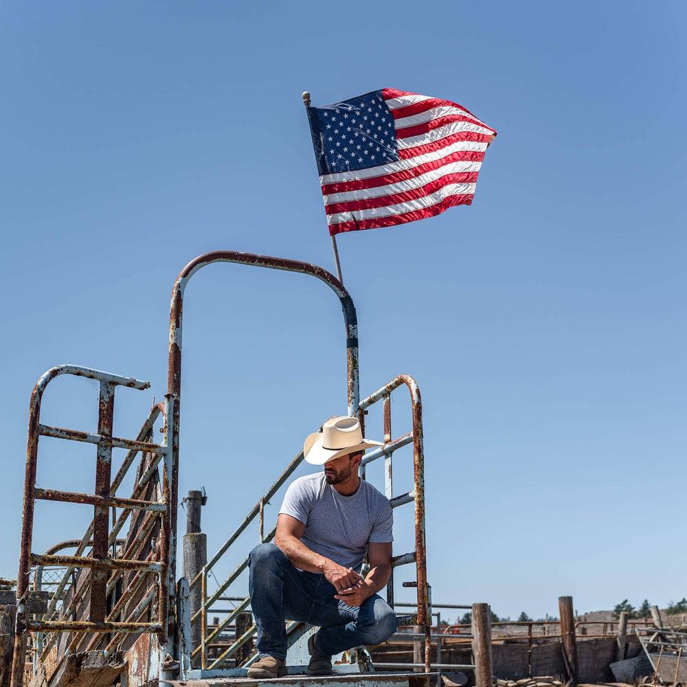 A man squatting on a metal stairway with US flag above him and wearing Cream Straw Cowboy Hat 