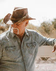 A man leaning on a wooden fence wearing a denim jacket and a cowboy hat